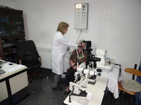 Doktorka u bijelom mantilu obavlja pregled oka pacijentu na aparatu, tabla sa optotipima u pozadini